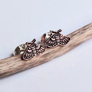 Pair of copper moth stud earrings with intricate moth detailing on a rustic wood surface