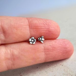 Tiny silver bee and flower mismatched stud earrings shown on fingertips for scale
