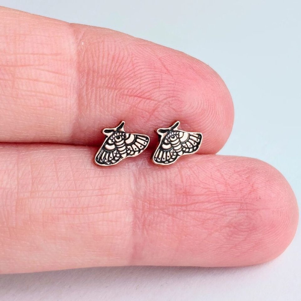 Small copper moth stud earrings held between fingertips to show their size